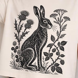 May include: A cream-colored short-sleeve t-shirt featuring a black ink-style graphic print of a rabbit sitting among thistles, clover, and blackberry branches. The design is centered on the front of the shirt, showcasing detailed botanical elements.