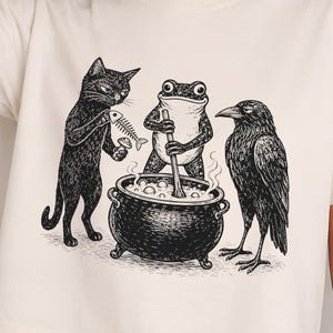 May include: A cream-colored short-sleeve t-shirt featuring a black ink-style graphic of a cat, a frog, and a raven gathered around a steaming cauldron. The cat holds a fish skeleton and a mushroom, while the frog stirs the pot with a wooden spoon.