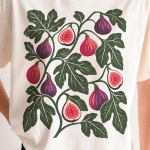 May include: A cream-colored short-sleeve t-shirt featuring a central graphic print of dark purple and red figs with green leaves. The illustration displays both whole figs and halved figs with visible seeds, arranged in a botanical pattern on the front.