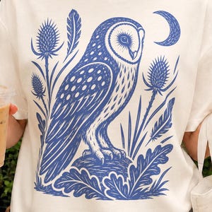 PNG design featuring a poised barn owl framed by teasel, oak leaves, scattered feathers and a crescent moon in a vintage block-print style in blue.