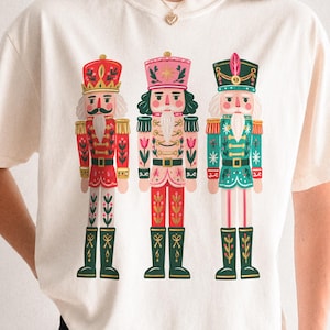 May include: A cream-colored short-sleeve t-shirt featuring a graphic print of three nutcracker figures standing side-by-side. The nutcrackers are illustrated in red, pink, and teal colors with gold accents, wearing tall hats and uniforms.