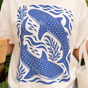 PNG design featuring two blue whale sharks swimming through stylized waves, seaweed, and small schools of fish in a bold linocut-inspired ocean scene.