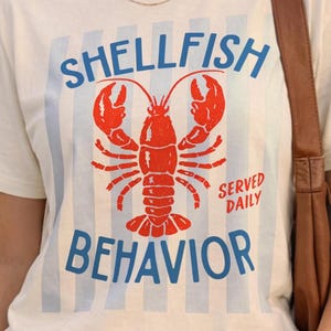 May include: A cream-colored short-sleeve t-shirt featuring a central graphic of a red lobster. The text reads "SHELLFISH BEHAVIOR" in blue letters above and below the lobster, with "SERVED DAILY" in red. The background has faint blue vertical stripes.