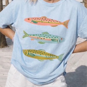 May include: A light blue short-sleeve t-shirt featuring a graphic print of three fish stacked vertically. The top fish is pink with green spots, the middle fish is light blue with green spots, and the bottom fish is olive green with dark spots.