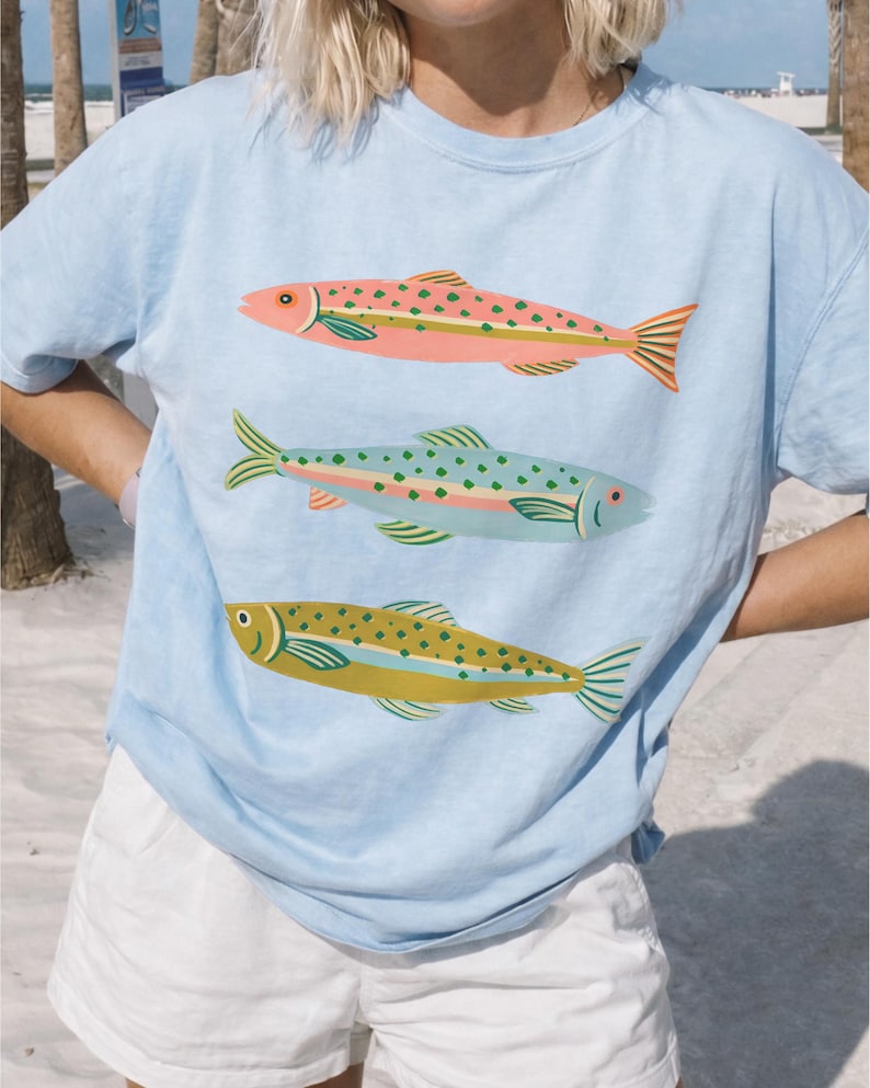 May include: A light blue short-sleeve t-shirt featuring a graphic print of three fish stacked vertically. The top fish is pink with green spots, the middle fish is light blue with green spots, and the bottom fish is olive green with dark spots.
