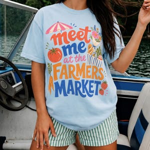 May include: A light blue t-shirt featuring a graphic print with the text "meet me at the FARMERS MARKET" in orange and blue. The design includes illustrations of a pink umbrella, a red tomato, orange carrots, strawberries, and a tote bag.
