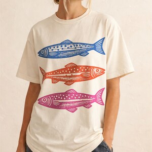 May include: Cream-colored t-shirt featuring three stylized fish in blue, orange, and pink. The fish are arranged vertically, each with a unique pattern of dots and lines. The shirt has short sleeves and a relaxed fit.