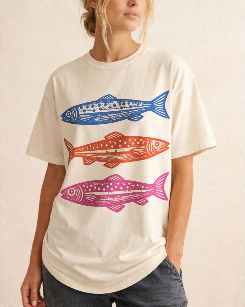 May include: Cream-colored t-shirt featuring three stylized fish in blue, orange, and pink. The fish are arranged vertically, each with a unique pattern of dots and lines. The shirt has short sleeves and a relaxed fit.