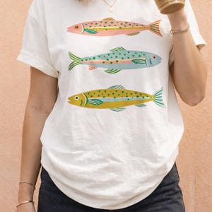 May include: A white short-sleeve t-shirt featuring a graphic print of three fish stacked vertically. The top fish is pink with green spots, the middle fish is light blue with green spots, and the bottom fish is yellow with green spots.