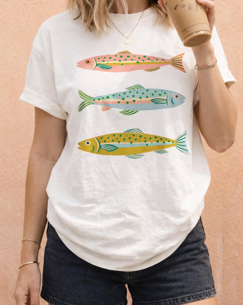 May include: A white short-sleeve t-shirt featuring a graphic print of three fish stacked vertically. The top fish is pink with green spots, the middle fish is light blue with green spots, and the bottom fish is yellow with green spots.