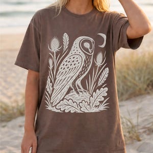 May include: A brown short-sleeve t-shirt featuring a large white graphic print of an owl perched on a rock, surrounded by thistle plants, leaves, feathers, and a crescent moon. The shirt has a crew neckline and a relaxed, loose-fitting silhouette.