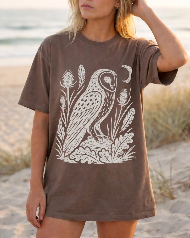 May include: A brown short-sleeve t-shirt featuring a large white graphic print of an owl perched on a rock, surrounded by thistle plants, leaves, feathers, and a crescent moon. The shirt has a crew neckline and a relaxed, loose-fitting silhouette.