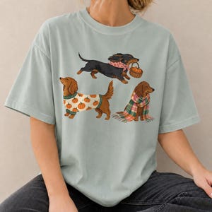May include: A sage green t-shirt featuring a graphic print of three dachshunds. One black dog wears a pink bandana and carries a basket, one brown dog wears a white sweater with orange pumpkins, and one brown dog is wrapped in a plaid scarf.