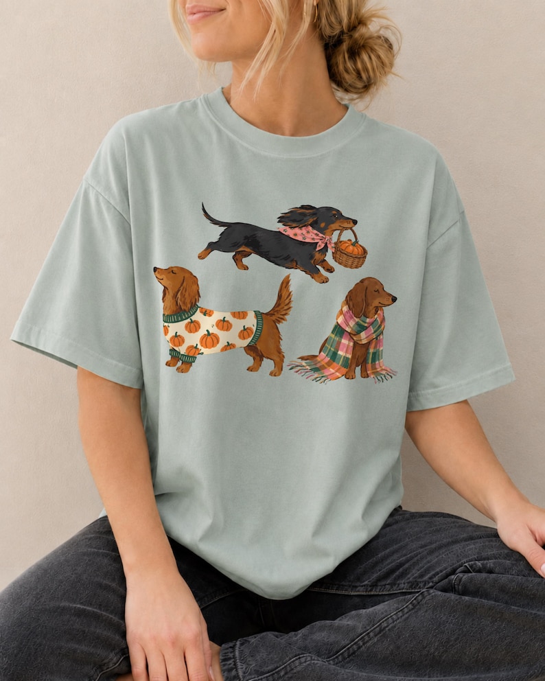 May include: A sage green t-shirt featuring a graphic print of three dachshunds. One black dog wears a pink bandana and carries a basket, one brown dog wears a white sweater with orange pumpkins, and one brown dog is wrapped in a plaid scarf.