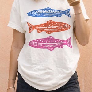 May include: White t-shirt featuring three stylized fish in blue, orange, and pink. The fish are arranged vertically, with detailed linework and dot patterns. The shirt has short sleeves and a relaxed fit, suitable for casual wear.