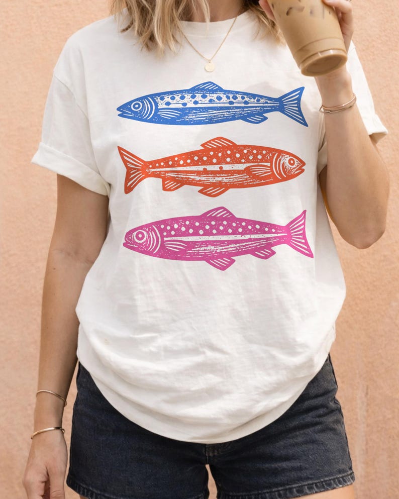 May include: White t-shirt featuring three stylized fish in blue, orange, and pink. The fish are arranged vertically, with detailed linework and dot patterns. The shirt has short sleeves and a relaxed fit, suitable for casual wear.