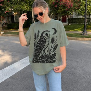 May include: An olive green short-sleeve t-shirt featuring a black graphic print of an owl perched on a rock, surrounded by thistle plants and a crescent moon. The shirt has a crew neckline and a relaxed fit, paired with light blue denim jeans.