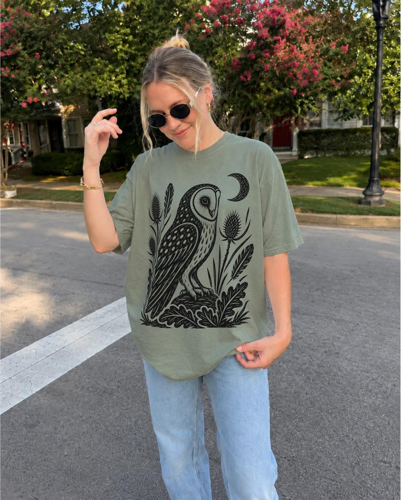 May include: An olive green short-sleeve t-shirt featuring a black graphic print of an owl perched on a rock, surrounded by thistle plants and a crescent moon. The shirt has a crew neckline and a relaxed fit, paired with light blue denim jeans.