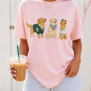 May include: A light pink short-sleeved t-shirt featuring a graphic print of three golden retriever dogs. The first dog wears a green sweater with a letter G, the second wears a blue floral bandana, and the third wears a white vest and green cap.