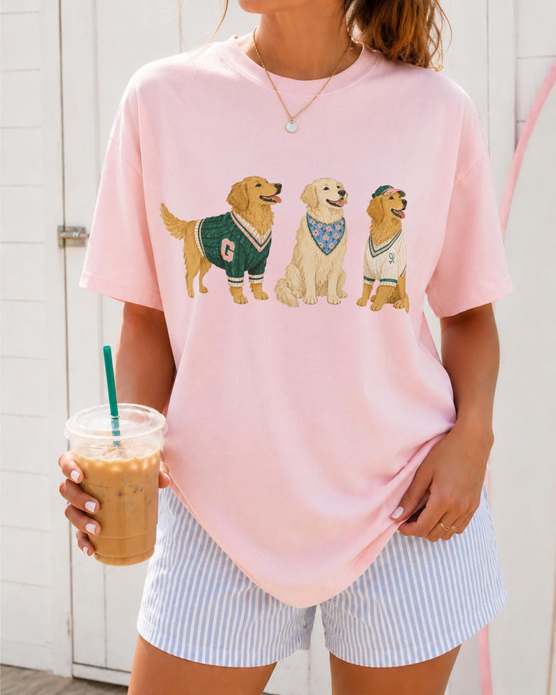May include: A light pink short-sleeved t-shirt featuring a graphic print of three golden retriever dogs. The first dog wears a green sweater with a letter G, the second wears a blue floral bandana, and the third wears a white vest and green cap.