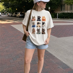 以下が含まれることがあります： オフホワイトの半袖Tシャツで、9匹の猫がグリッド状に並んだグラフィックプリントが施されています。それぞれの猫は、スカーフ、サングラス、ネックレス、セーターなど、さまざまな色や柄のアクセサリーを身につけて描かれています。