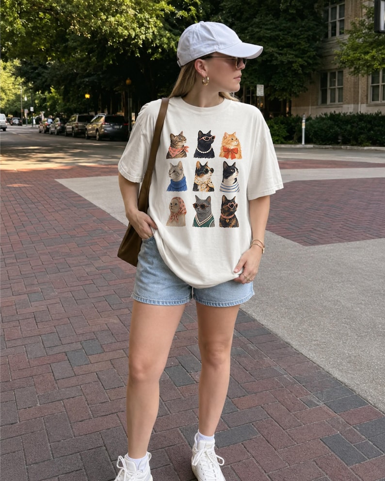 以下が含まれることがあります： オフホワイトの半袖Tシャツで、9匹の猫がグリッド状に並んだグラフィックプリントが施されています。それぞれの猫は、スカーフ、サングラス、ネックレス、セーターなど、さまざまな色や柄のアクセサリーを身につけて描かれています。