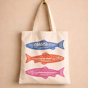 May include: Cream-colored tote bag with three colorful fish designs. The fish are blue, orange, and pink, each with a unique pattern. The bag hangs from a gold hook, showcasing a simple yet eye-catching design.