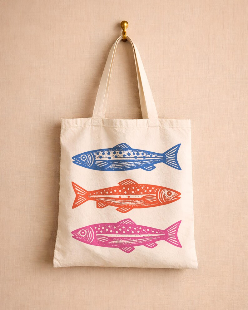 May include: Cream-colored tote bag with three colorful fish designs. The fish are blue, orange, and pink, each with a unique pattern. The bag hangs from a gold hook, showcasing a simple yet eye-catching design.