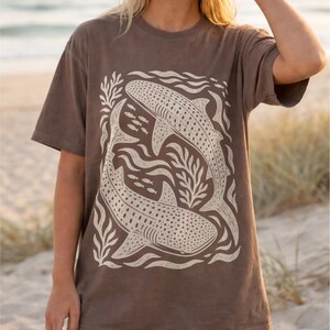Op de afbeelding: Een bruin t-shirt met korte mouwen en een grote cr&egrave;mekleurige grafische print van twee walvishaaien die in een cirkelvormige beweging zwemmen, omringd door gestileerd zeewier en kleine vissen. Het ontwerp heeft een getextureerde blokdruk-look.