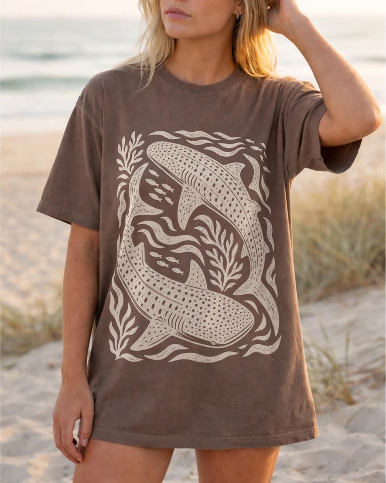 Op de afbeelding: Een bruin t-shirt met korte mouwen en een grote cr&egrave;mekleurige grafische print van twee walvishaaien die in een cirkelvormige beweging zwemmen, omringd door gestileerd zeewier en kleine vissen. Het ontwerp heeft een getextureerde blokdruk-look.