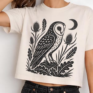 May include: A cream-colored short-sleeve t-shirt featuring a black graphic print of an owl perched on a rock, surrounded by thistle plants, feathers, and a crescent moon. The shirt has a crew neckline and a straight hemline at the waist.