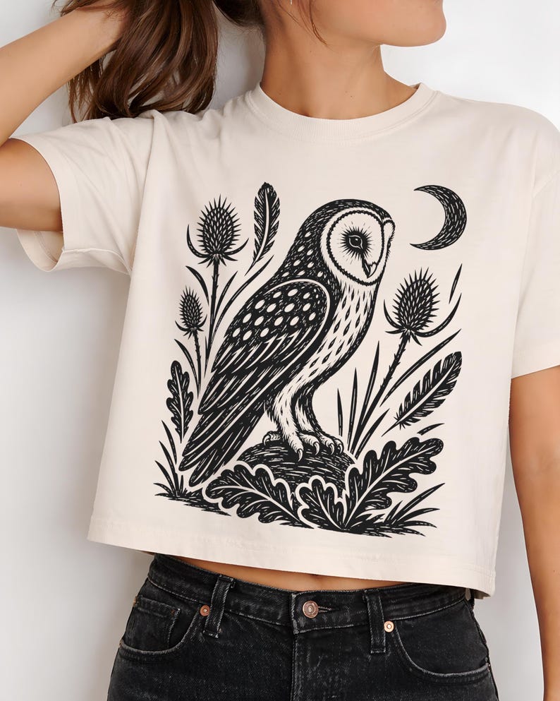 May include: A cream-colored short-sleeve t-shirt featuring a black graphic print of an owl perched on a rock, surrounded by thistle plants, feathers, and a crescent moon. The shirt has a crew neckline and a straight hemline at the waist.