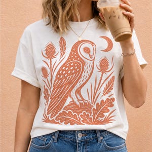 May include: A white short-sleeve t-shirt featuring a large, detailed orange graphic print of an owl perched on a mound, surrounded by stylized thistles, leaves, and a crescent moon. The shirt has a crew neckline and a relaxed, casual fit.