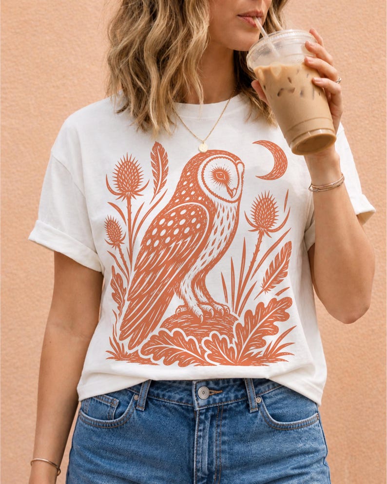May include: A white short-sleeve t-shirt featuring a large, detailed orange graphic print of an owl perched on a mound, surrounded by stylized thistles, leaves, and a crescent moon. The shirt has a crew neckline and a relaxed, casual fit.