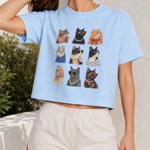 以下が含まれることがあります： 水色の半袖クロップトップで、9匹の猫がグリッド状に並んだグラフィックプリントが施されています。それぞれの猫は、スカーフ、サングラス、パールのネックレス、セーターなど、さまざまな色や柄の異なるアクセサリーを身につけています。