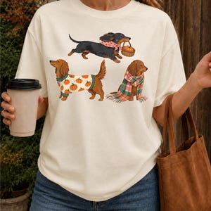 May include: An off-white short-sleeve t-shirt featuring a graphic print of three dachshunds. One dog wears a pink bandana and carries a pumpkin basket, another wears a pumpkin-patterned sweater, and the third is wrapped in a plaid scarf.