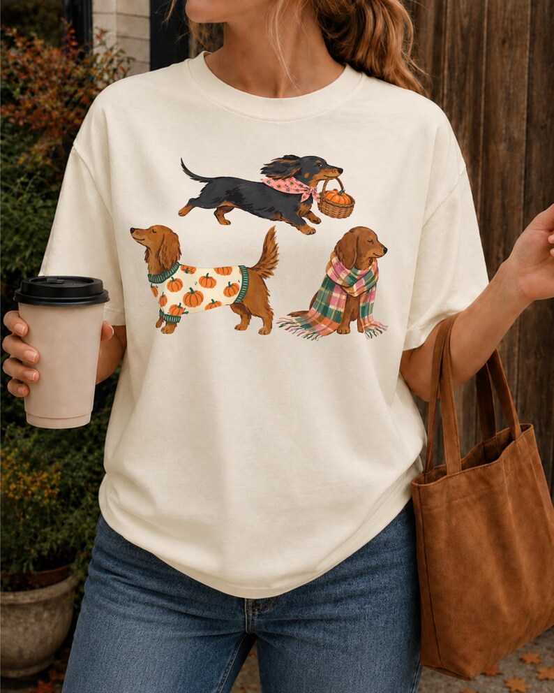 May include: An off-white short-sleeve t-shirt featuring a graphic print of three dachshunds. One dog wears a pink bandana and carries a pumpkin basket, another wears a pumpkin-patterned sweater, and the third is wrapped in a plaid scarf.