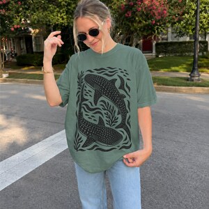 Op de afbeelding: Een saliegroen T-shirt met korte mouwen en een zwarte grafische print van twee walvishaaien die tussen gestileerd zeewier en kleine vissen zwemmen. Het T-shirt heeft een ronde hals en een relaxte pasvorm met een gedetailleerde illustratie.