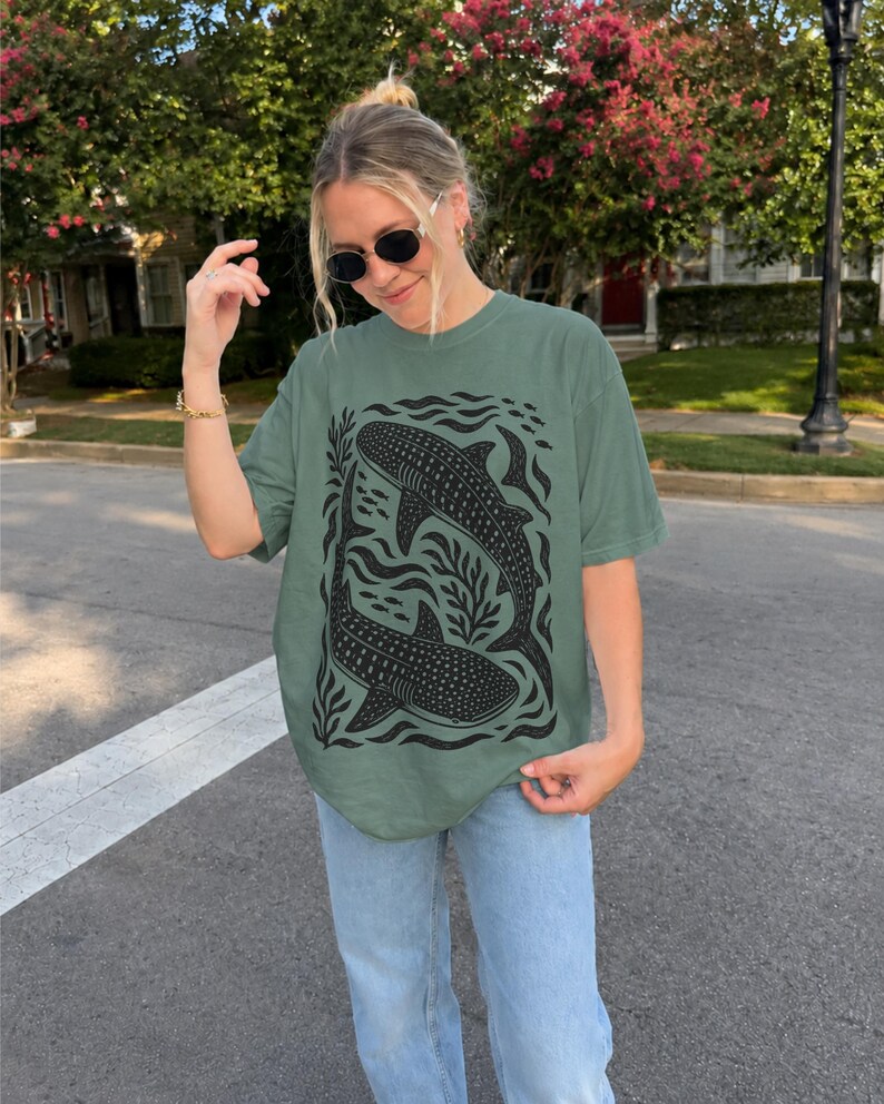 Op de afbeelding: Een saliegroen T-shirt met korte mouwen en een zwarte grafische print van twee walvishaaien die tussen gestileerd zeewier en kleine vissen zwemmen. Het T-shirt heeft een ronde hals en een relaxte pasvorm met een gedetailleerde illustratie.