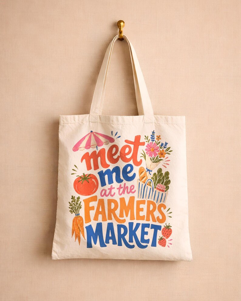 May include: A cream-colored canvas tote bag featuring a colorful graphic print with the text "meet me at the FARMERS MARKET". The design includes illustrations of a pink and white striped umbrella, a bouquet of flowers, a tomato, carrots, and strawberries.