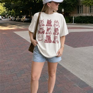 May include: An off-white short-sleeve t-shirt featuring a grid of nine maroon-colored cat illustrations, each wearing different accessories like glasses, scarves, or ties. The shirt is paired with light blue denim shorts, a white baseball cap, and a brown bag.
