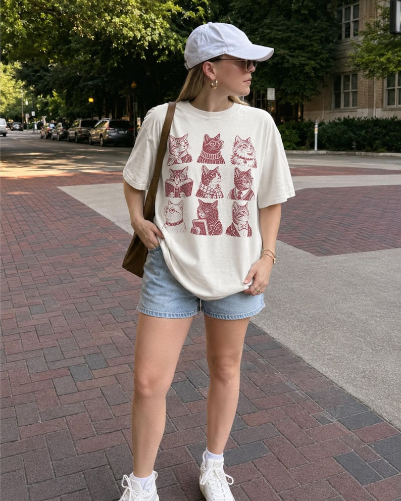 May include: An off-white short-sleeve t-shirt featuring a grid of nine maroon-colored cat illustrations, each wearing different accessories like glasses, scarves, or ties. The shirt is paired with light blue denim shorts, a white baseball cap, and a brown bag.