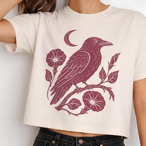 May include: A cream-colored short-sleeve crop top featuring a dark maroon graphic print of a crow perched on a flowering vine with morning glory blossoms and a crescent moon positioned above the bird, set against a plain light background.