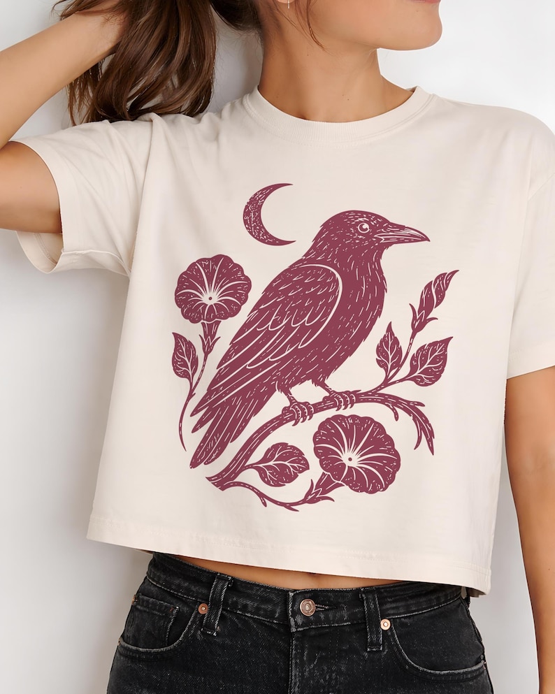 May include: A cream-colored short-sleeve crop top featuring a dark maroon graphic print of a crow perched on a flowering vine with morning glory blossoms and a crescent moon positioned above the bird, set against a plain light background.