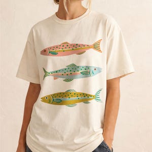 May include: An off-white short-sleeve t-shirt featuring a graphic print of three fish stacked vertically. The top fish is pink with green spots, the middle fish is light blue with green spots, and the bottom fish is yellow with green spots.