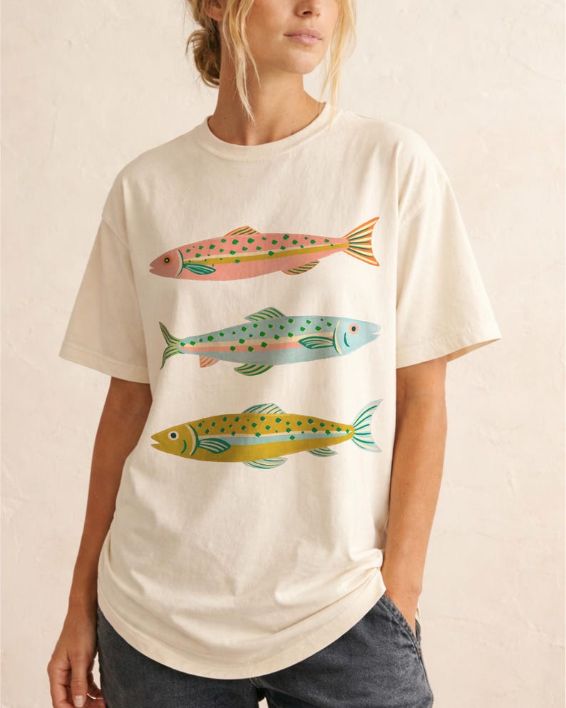 May include: An off-white short-sleeve t-shirt featuring a graphic print of three fish stacked vertically. The top fish is pink with green spots, the middle fish is light blue with green spots, and the bottom fish is yellow with green spots.