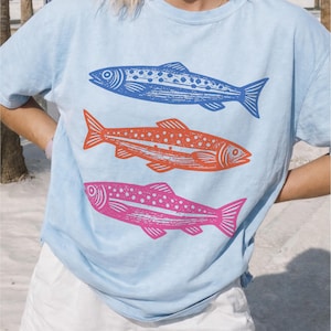 May include: Light blue t-shirt featuring three stylized fish in blue, orange, and pink. The fish are arranged vertically, with detailed scales and fins. The shirt is a casual style, suitable for everyday wear.