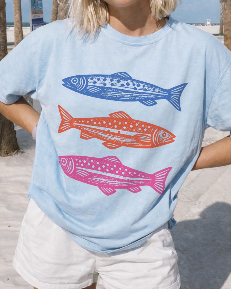May include: Light blue t-shirt featuring three stylized fish in blue, orange, and pink. The fish are arranged vertically, with detailed scales and fins. The shirt is a casual style, suitable for everyday wear.
