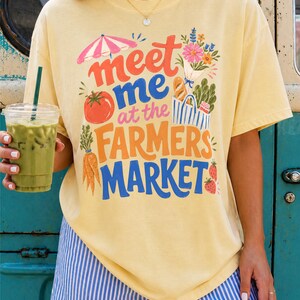 May include: A pale yellow short-sleeve t-shirt featuring a colorful graphic print with the text "meet me at the FARMERS MARKET" in red, blue, and orange. The design includes illustrations of a pink umbrella, flowers, a tomato, carrots, and strawberries.
