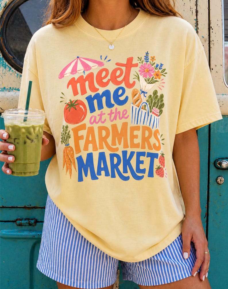 May include: A pale yellow short-sleeve t-shirt featuring a colorful graphic print with the text "meet me at the FARMERS MARKET" in red, blue, and orange. The design includes illustrations of a pink umbrella, flowers, a tomato, carrots, and strawberries.